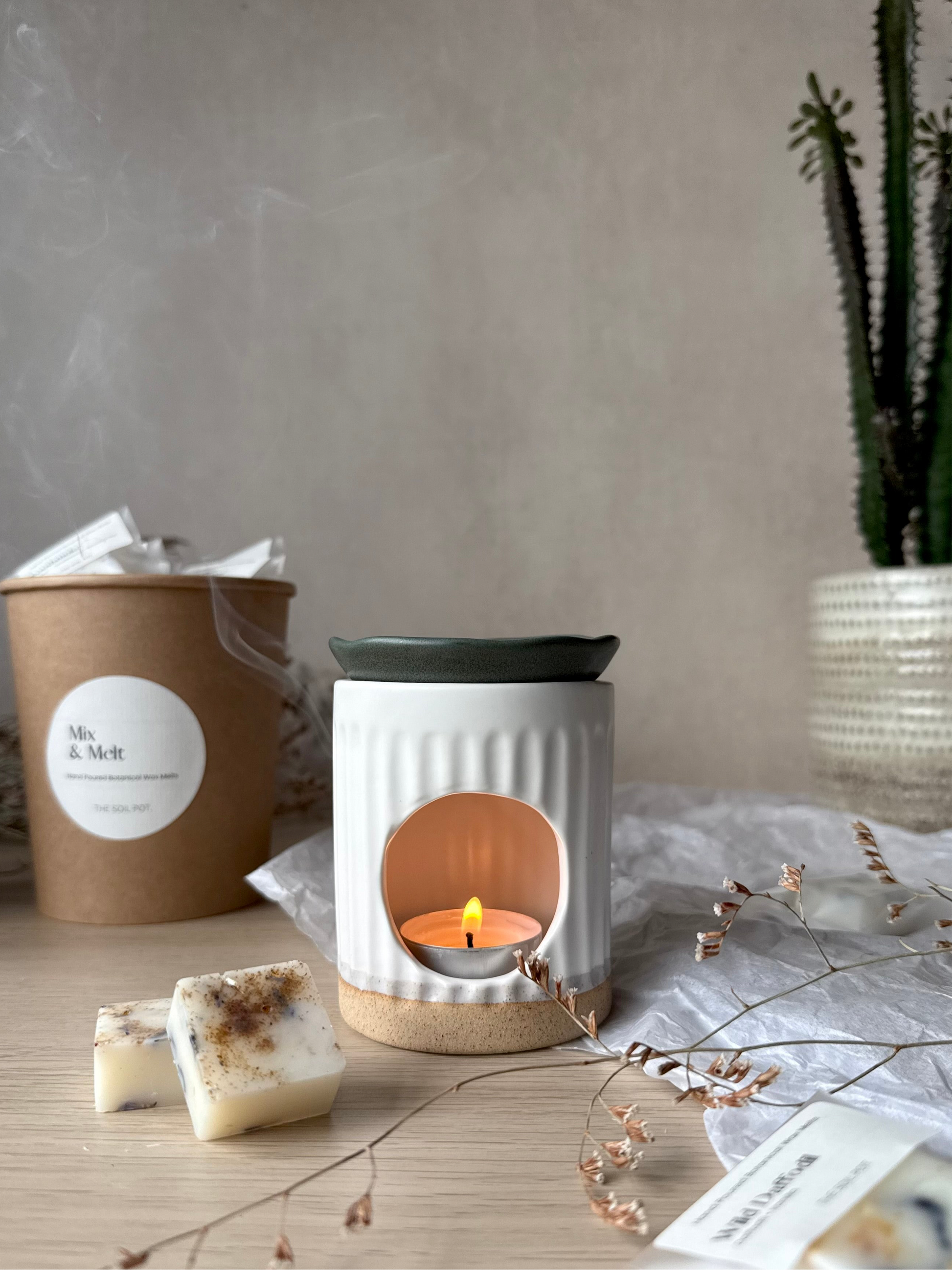White wax warmer with a lit candle on a wooden surface, surrounded by wax melts.