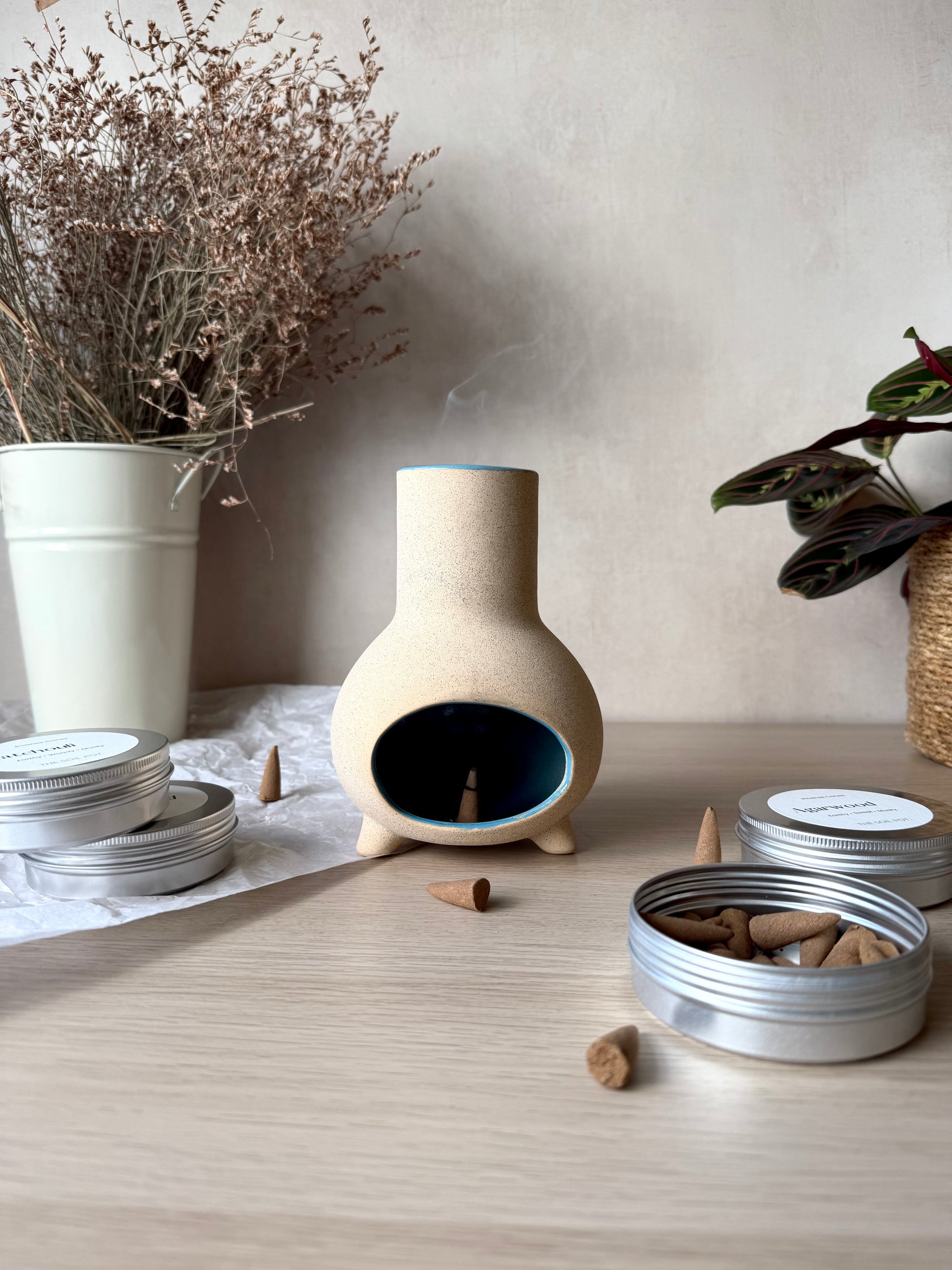 Ceramic incense burner with incense cones on a wooden surface, surrounded by plants.