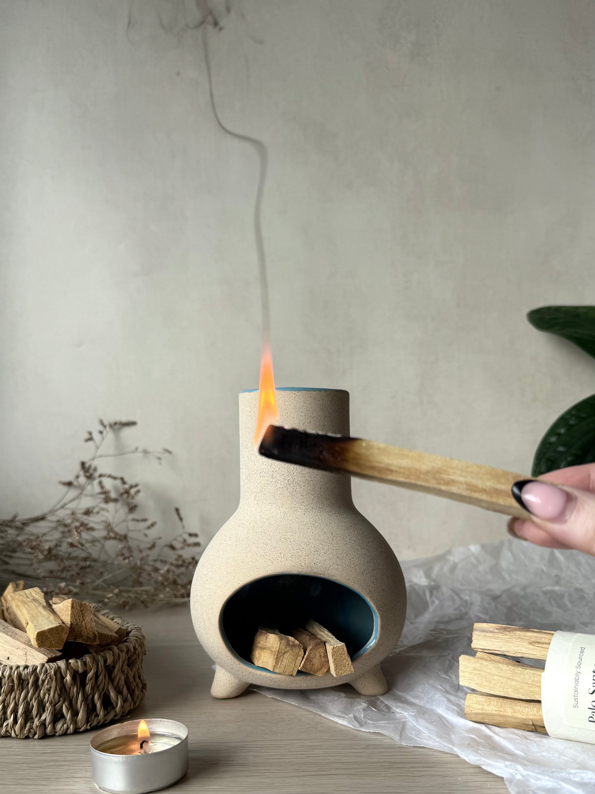 Person lighting palo santo in front of a mini chiminea burner, on a neutral background.