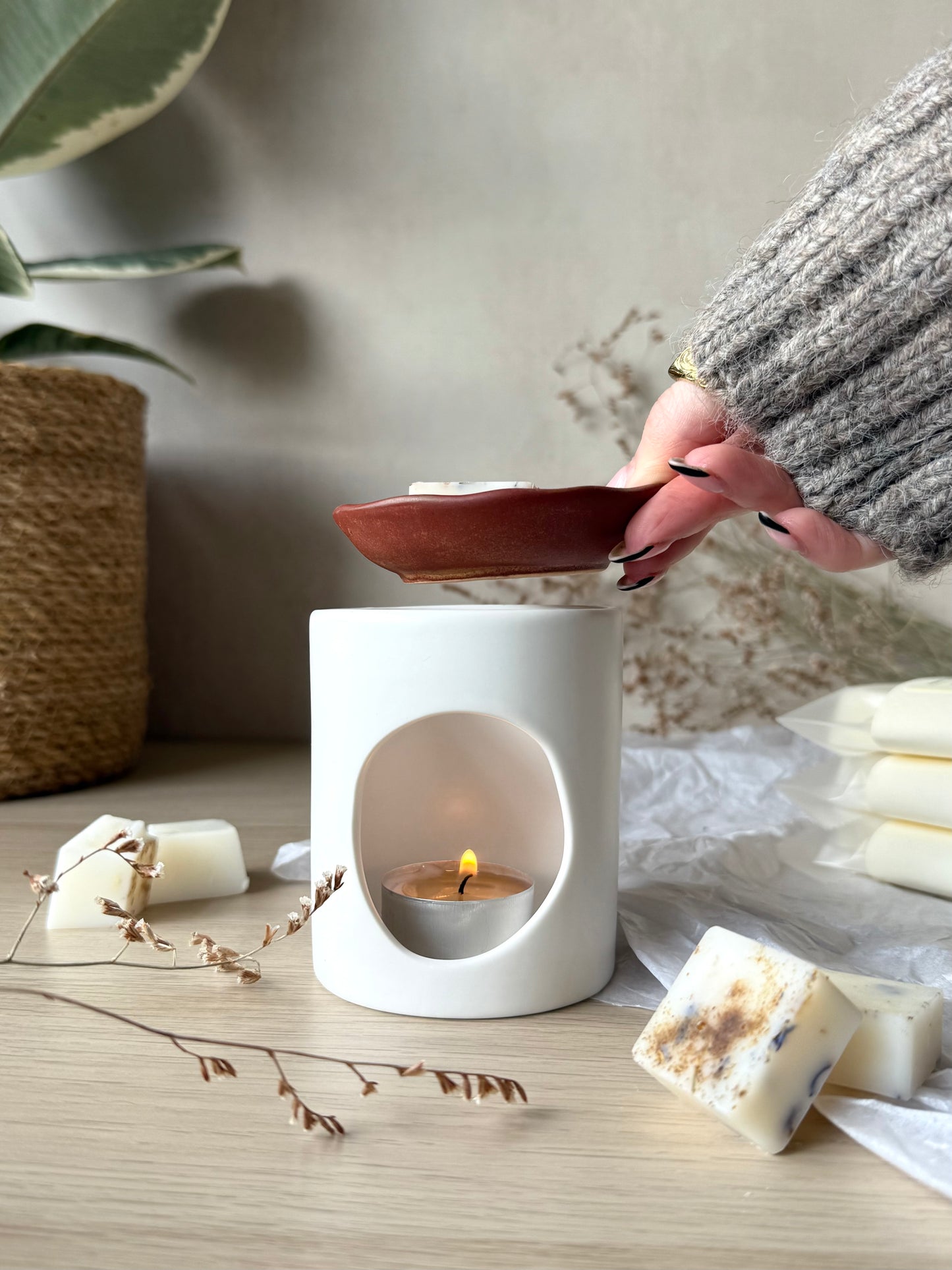 Person using a white wax melter with a lit candle on a wooden surface, surrounded by wax bars and a plant.